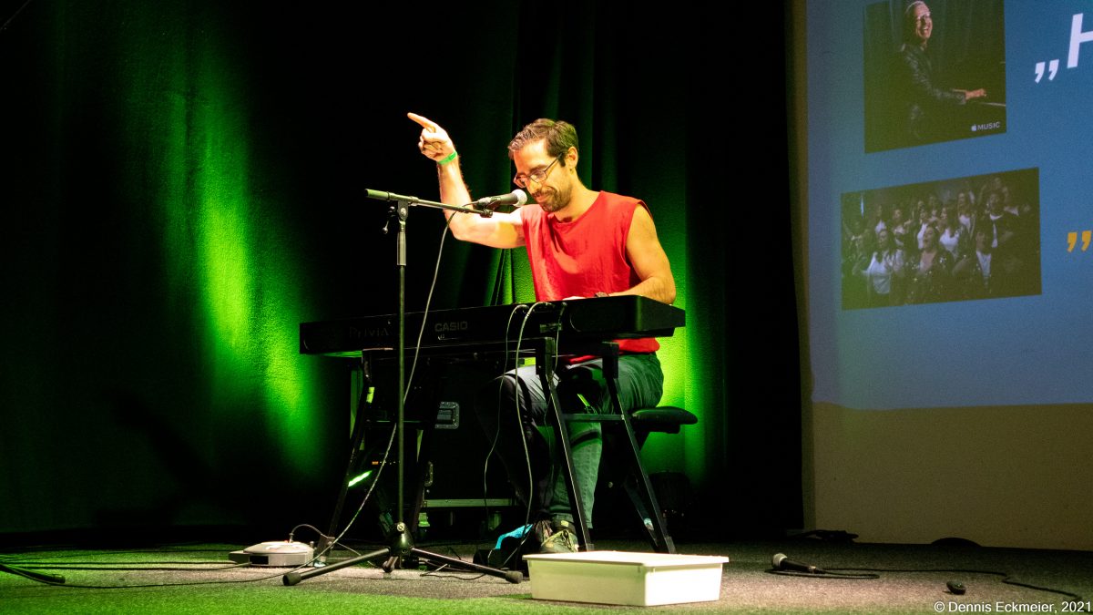 Slammer an elektronischem Klavier. Er zeigt auf das Publikum. Im Hintergrund ist seine Präsentation auf der Leinwand.