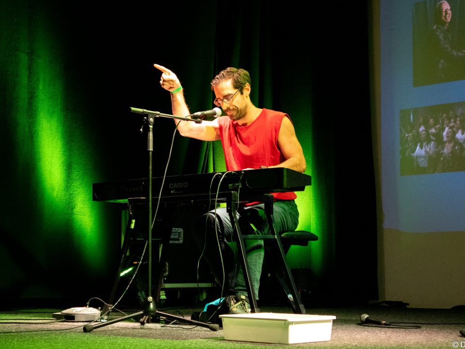 Slammer an elektronischem Klavier. Er zeigt auf das Publikum. Im Hintergrund ist seine Präsentation auf der Leinwand.