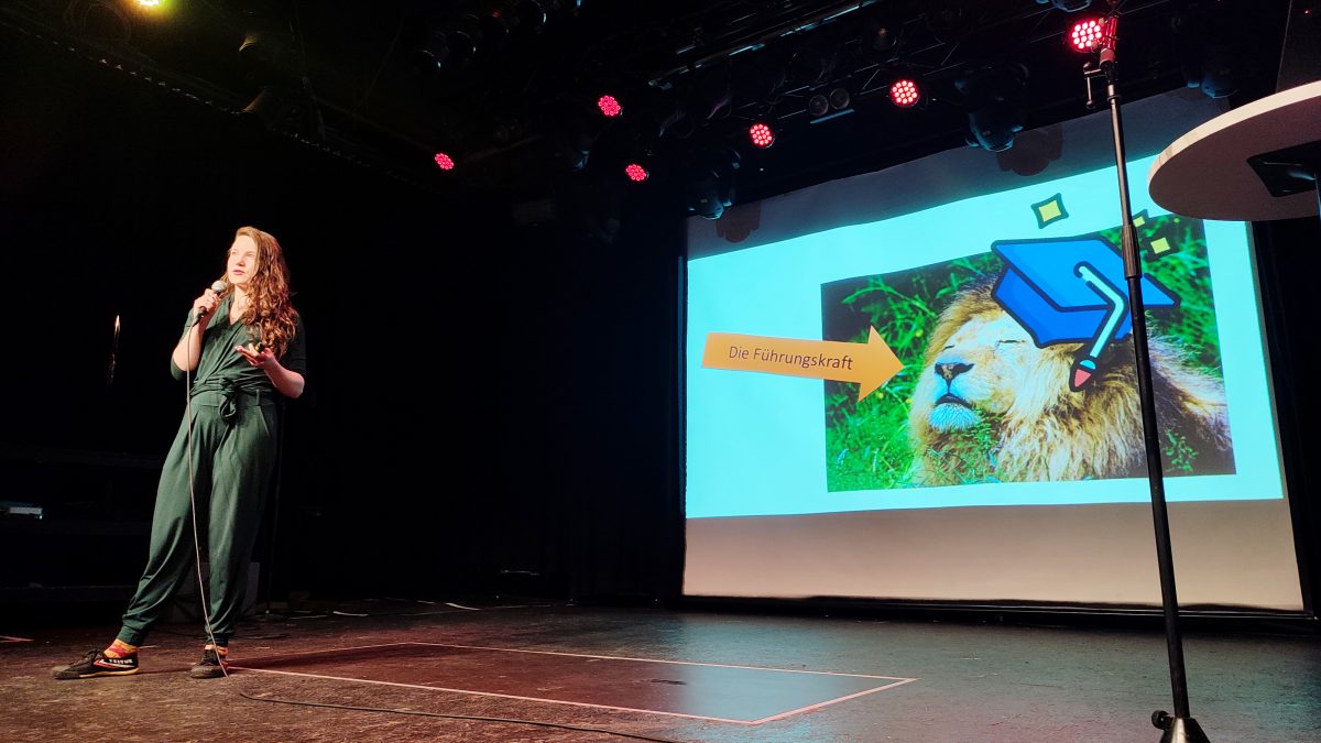 Linn Voss hält ihren Science-Slam-Vortrag auf der Bühne. Das aktulle Slide zeigt einen Löwen der als Führungskraft dargestellt wird,