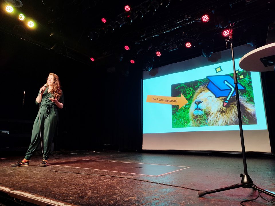 Linn Voss hält ihren Science-Slam-Vortrag auf der Bühne. Das aktulle Slide zeigt einen Löwen der als Führungskraft dargestellt wird,