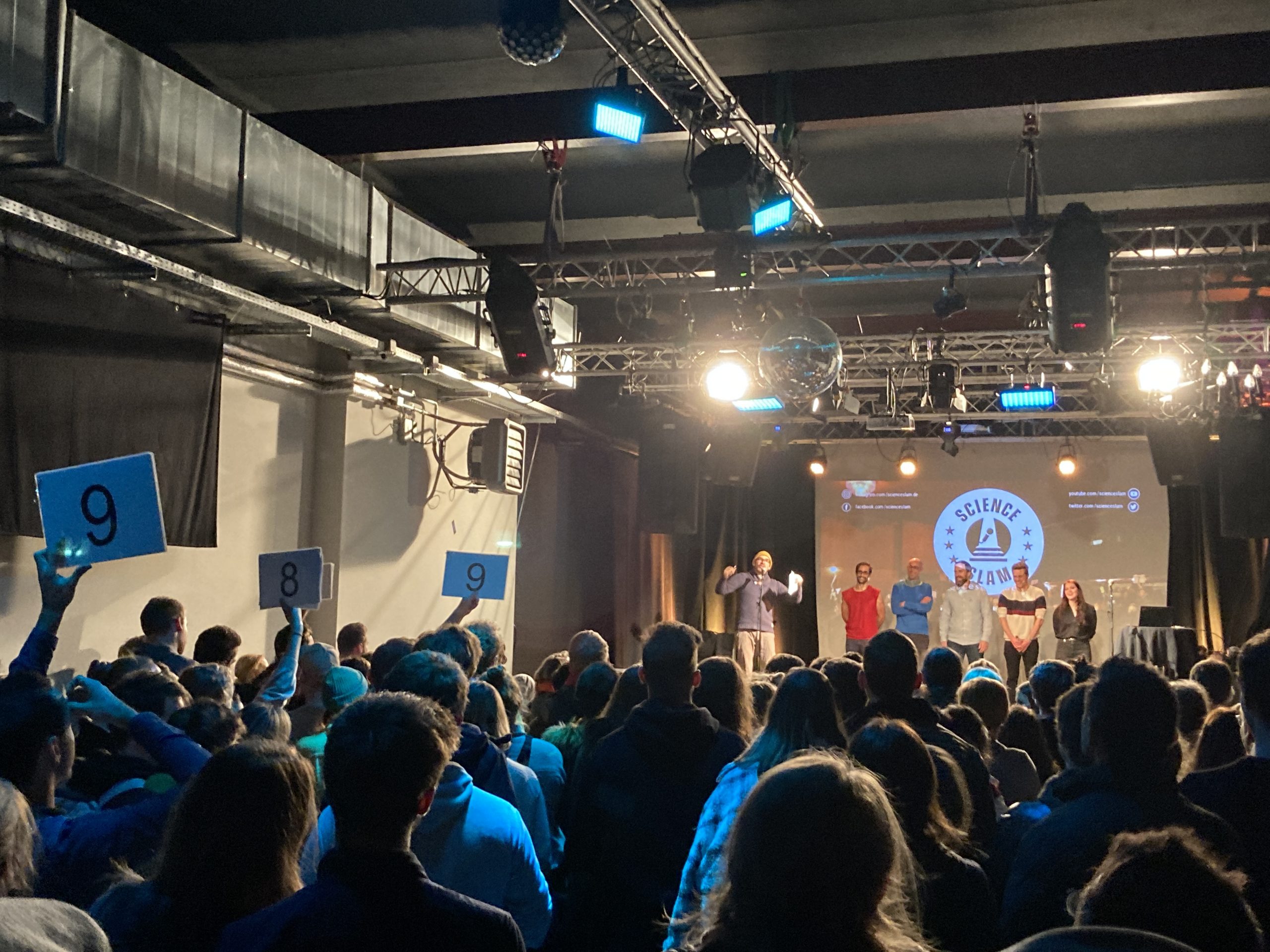 Publikum vor der Bühne eines Science Slams hält die Wertungstafeln hoch