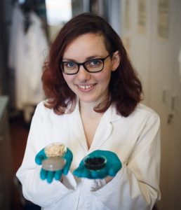 Maria-Elena Vorrath ist in die Kamera lächelnd zu sehen. Sie trägt einen weißen Laborkittel und blaue Laborhandschuhe. In den Händen hält sie Petrischalen mit Proben ihrer Arbeit.